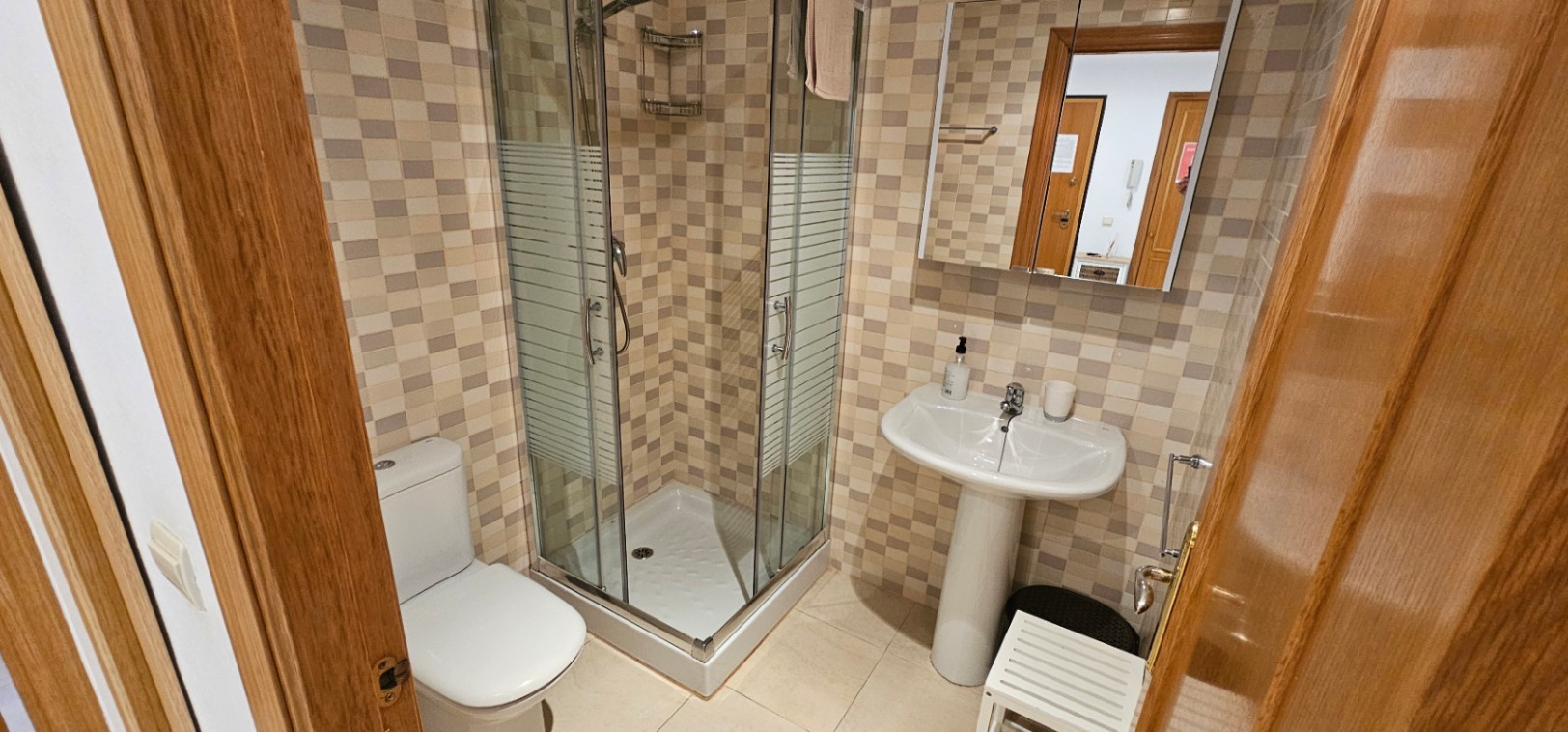 Funktionales Badezimmer mit einer Eckglasdusche, einem Säulenwaschbecken und dekorativen Mosaikwandfliesen in warmen Erdtönen.