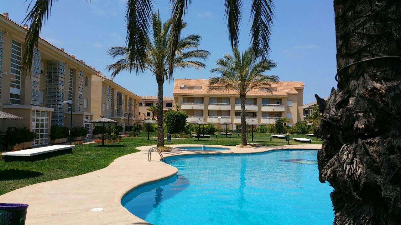 Gemeinschaftsanlage mit großem, geschwungenem Pool, gepflegten Rasenflächen und hohen Palmen vor den mediterranen Apartmentgebäuden.
