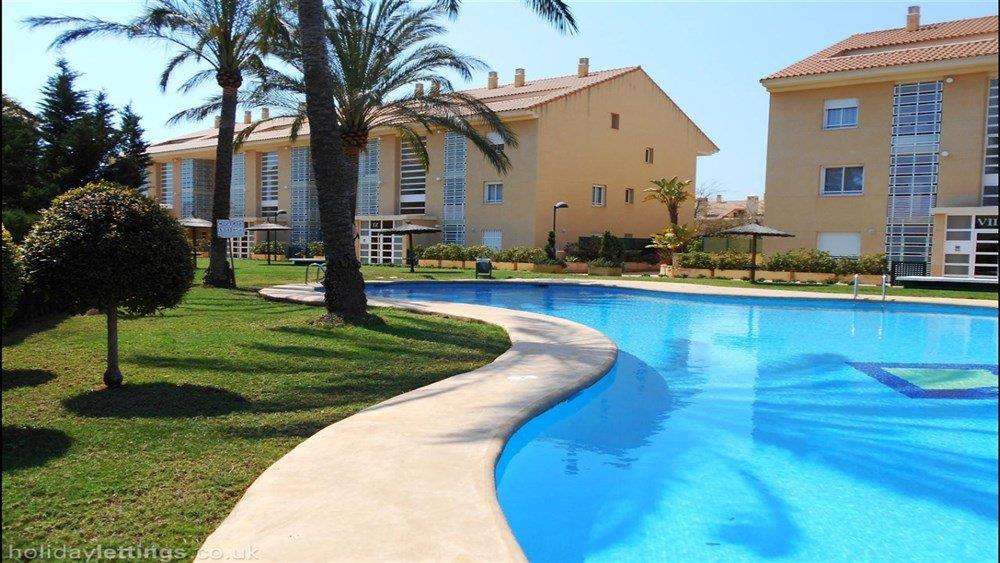 Großer Gemeinschaftspool mit geschwungenen Formen, eingebettet in eine gepflegte Gartenanlage mit Palmen und mediterranen Wohngebäuden.