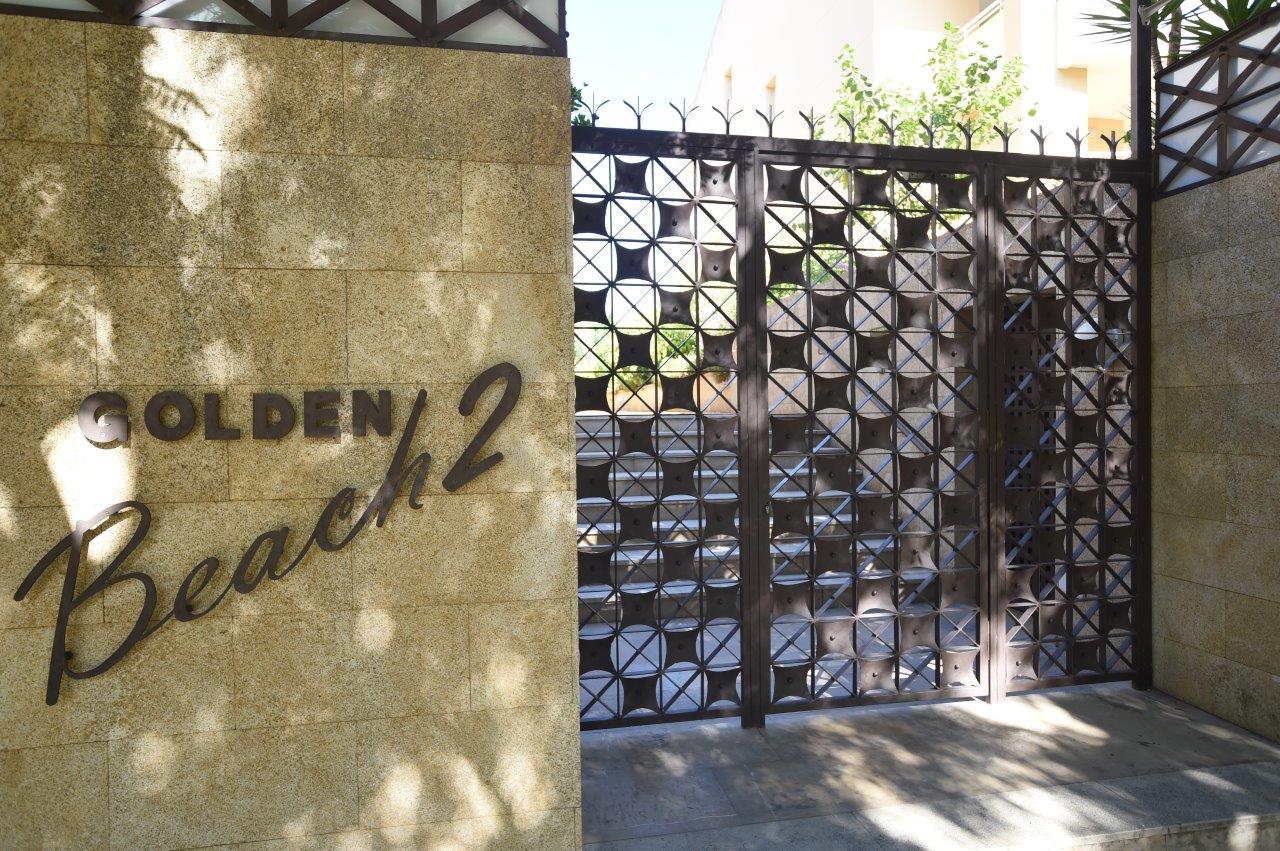 Haupteingang der Anlage Golden Beach 2 mit Natursteinmauer, Metallbeschriftung und einem dekorativen Sicherheitstor mit geometrischem Muster.