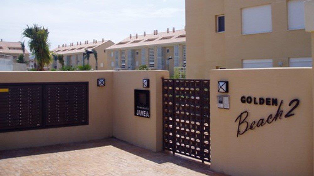 Haupteingang der Wohnanlage Golden Beach 2 in Javea mit beiger Umfassungsmauer, dunklem Metalltor und integrierten Briefkästen.