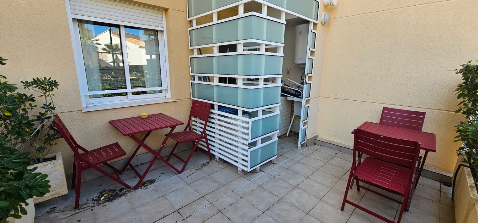 Private geflieste Terrasse mit weinroten Gartenmöbeln und einer funktionalen Abtrennung aus weißen Lamellen und Milchglas für den Hauswirtschaftsbereich.