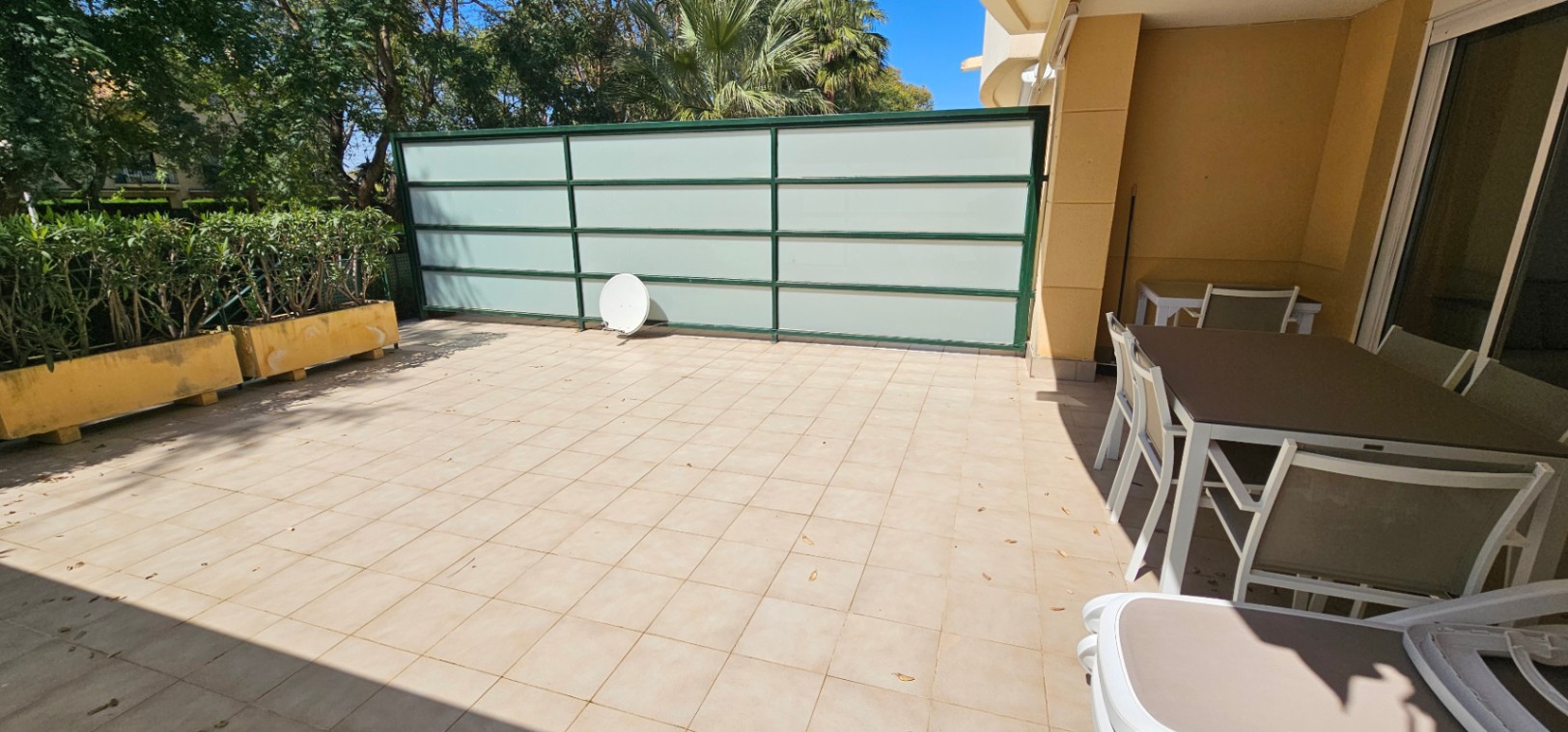 Private Südterrasse mit Fliesenboden, Essbereich im Freien und Sichtschutzwänden, umgeben von Pflanzgefäßen und altem Baumbestand.