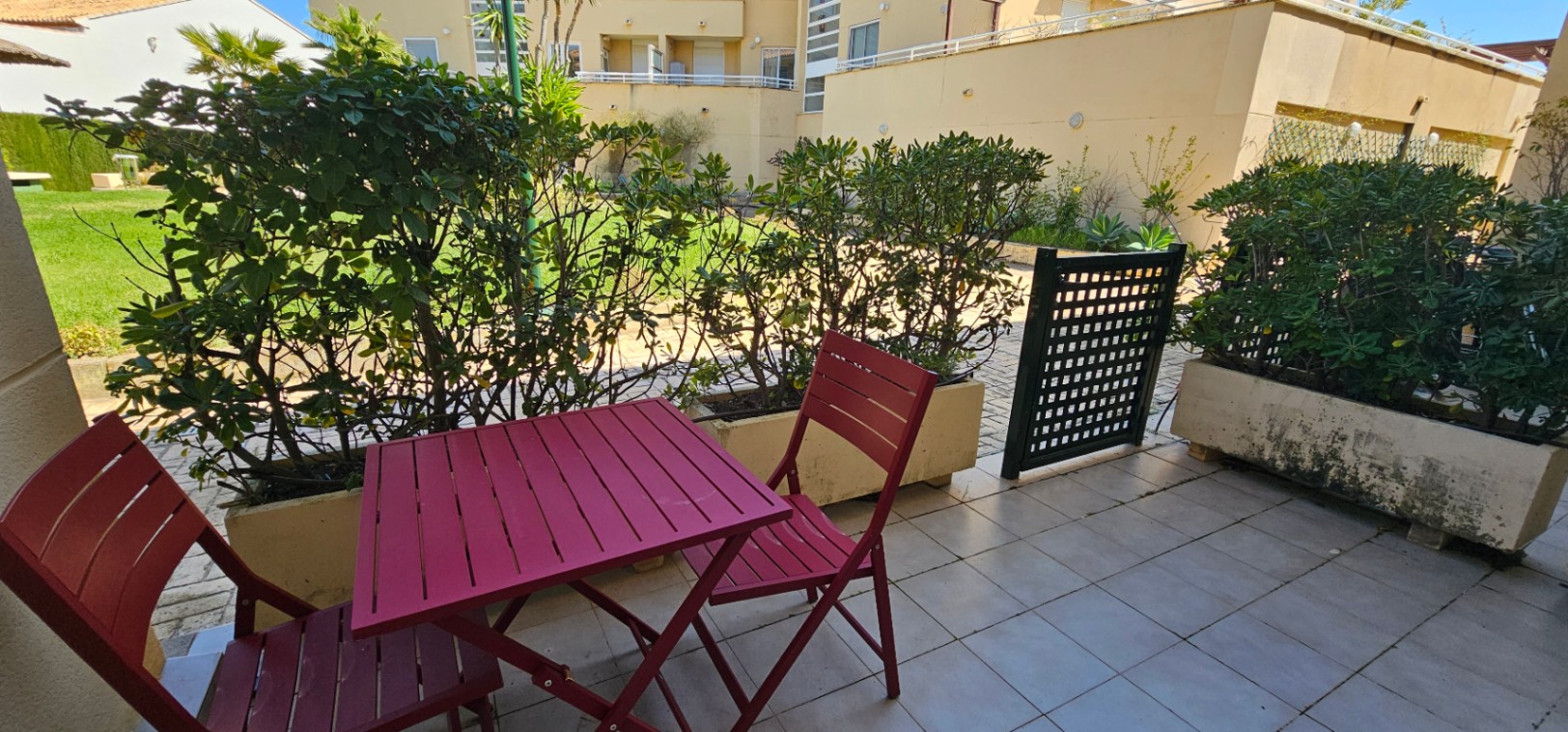 Private Terrasse mit hellen Bodenfliesen, burgunderroten Metall-Gartenmöbeln und direktem Zugang zu den gemeinschaftlichen Grünanlagen.