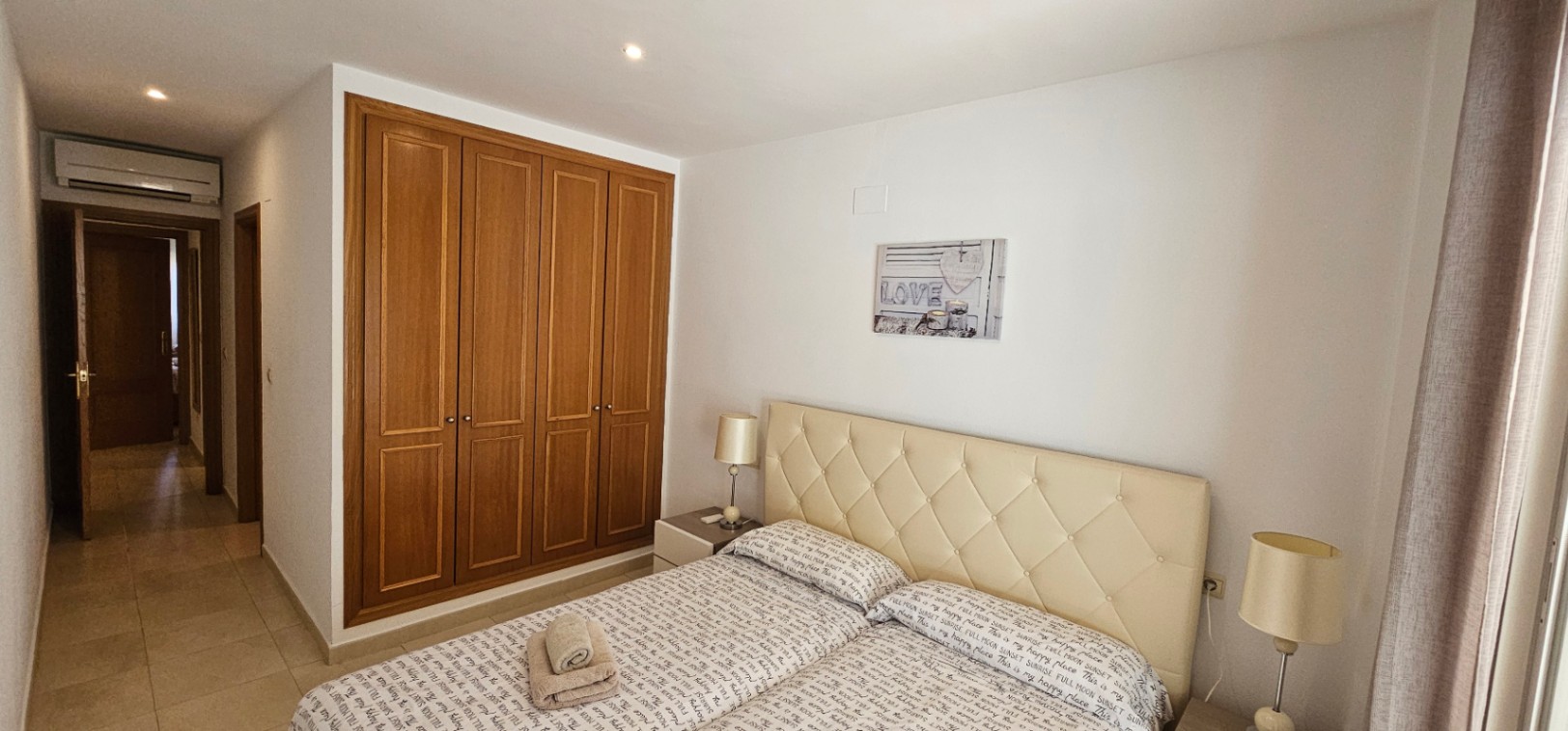 Schlafzimmer mit vierteiligem Einbauschrank aus Holz, cremefarbenem, gepolstertem Kopfteil und Klimaanlage über dem Flurdurchgang.
