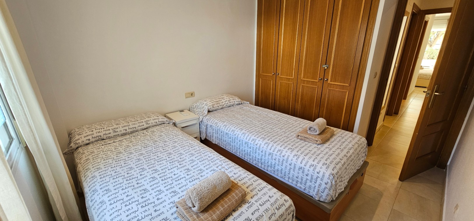 Schlafzimmer mit zwei Einzelbetten, einem großen viertürigen Holzeinbauschrank und hellen Fliesenböden mit Zugang zum Flur.