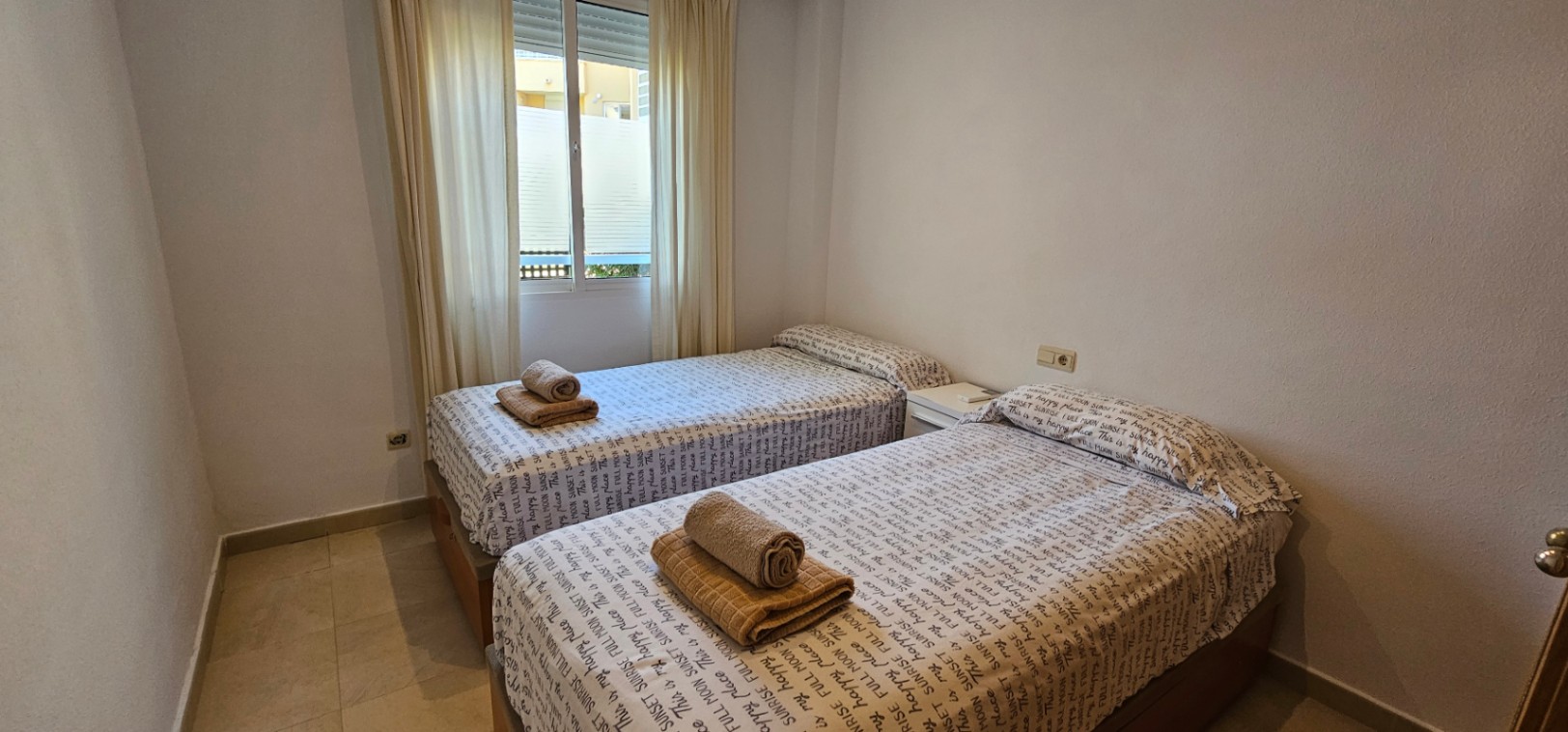 Schlafzimmer mit zwei Einzelbetten, hellem Fliesenboden und einem Fenster mit weißen Vorhängen für natürliches Licht.