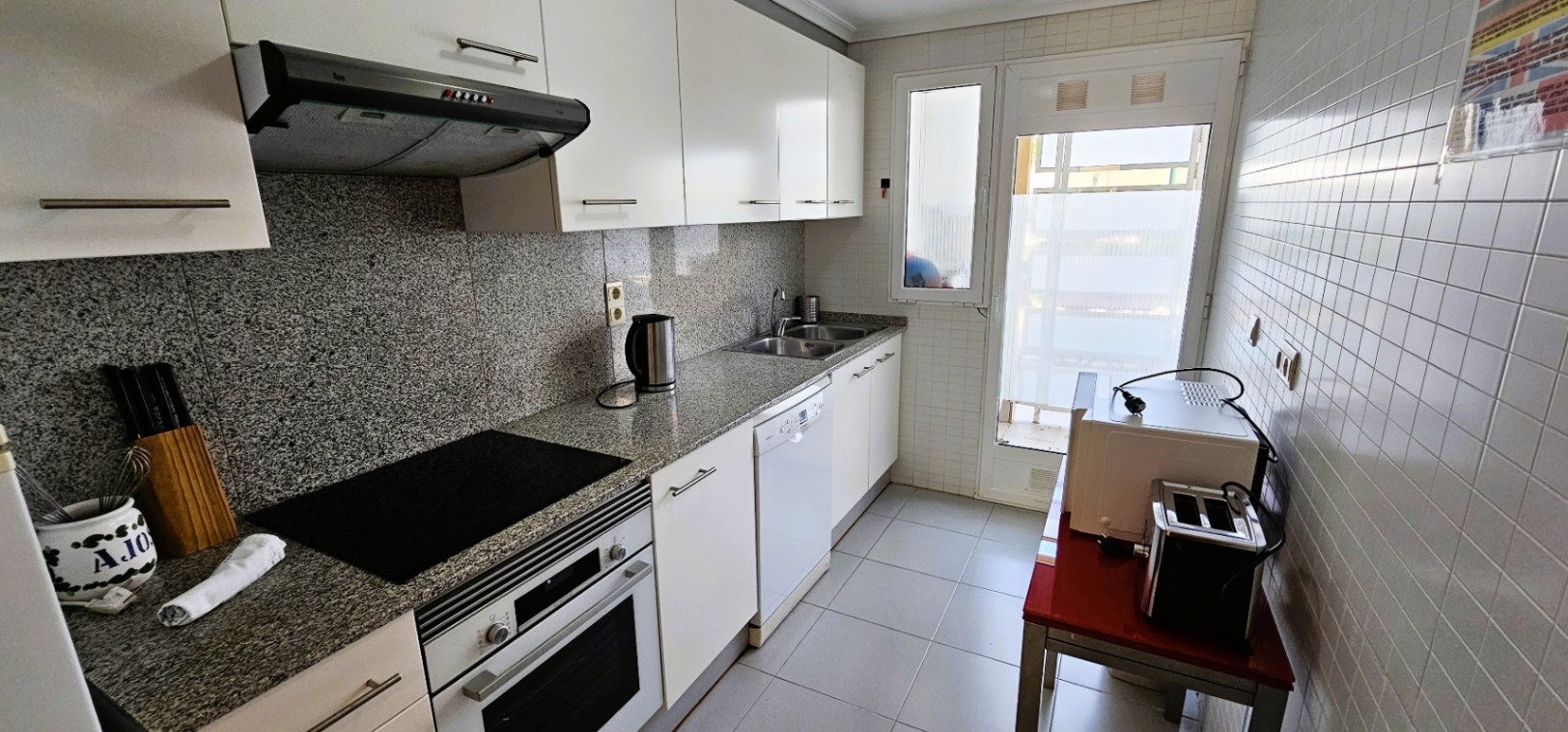 Separate Küche mit Granitarbeitsplatte, weißen Schränken, integriertem Backofen, Cerankochfeld und Glastür für Tageslicht und Zugang nach draußen.