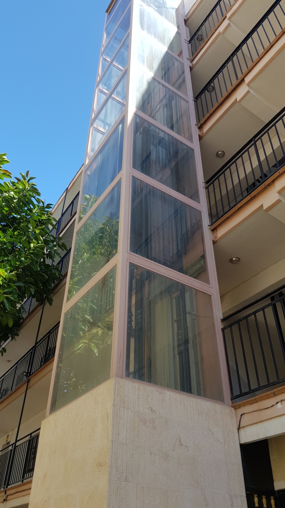 Außenliegender Panorama-Glasaufzug mit Steinsockel, integriert in die Fassade des Wohngebäudes neben Balkonen mit schwarzen Metallgeländern.