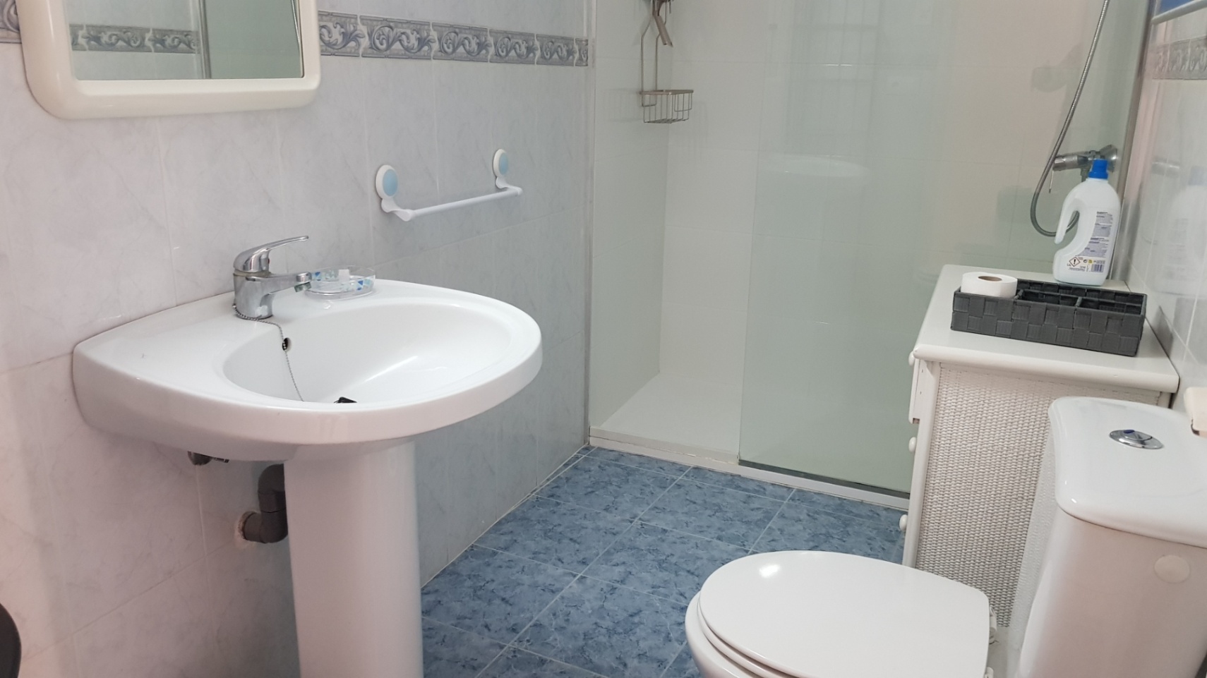 Badezimmer mit blauen Bodenfliesen, Standwaschbecken und Dusche mit Glastrennwand. Weiße Wandfliesen mit dekorativer Bordüre sorgen für ein helles Ambiente.