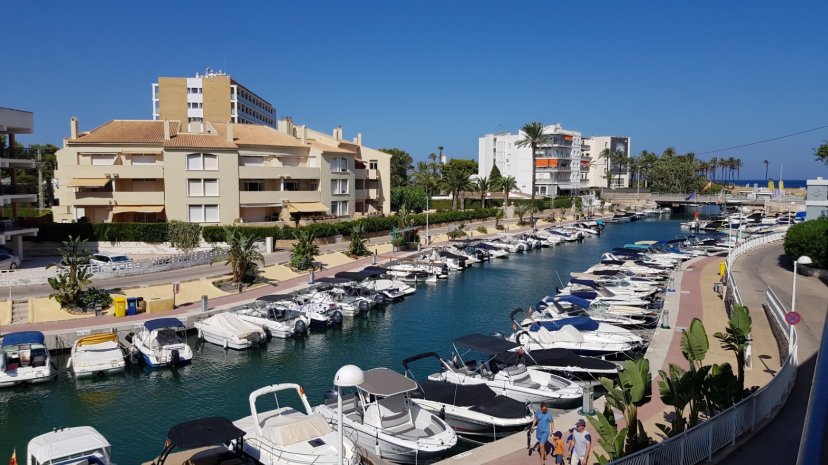 Erhöhter Blick von der Terrasse auf den Jachthafenkanal mit angelegten Freizeitbooten, mediterranen Gebäuden und Palmen bei klarem Himmel.