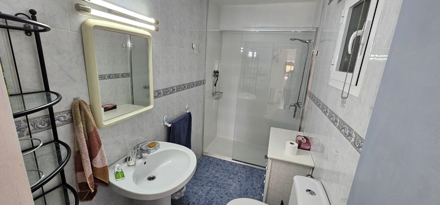 Funktionales Badezimmer mit begehbarer Dusche, Glastrennwand, Säulenwaschbecken und blauen Bodenfliesen, ergänzt durch dekorative Wandbordüren.