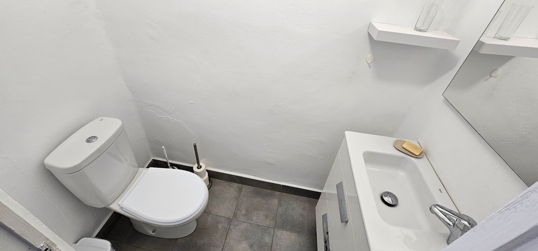 Funktionales Badezimmer mit weißem WC, Waschtisch mit integriertem Waschbecken und grauen Bodenfliesen. Inklusive Spiegel und minimalistischem Wandregal.