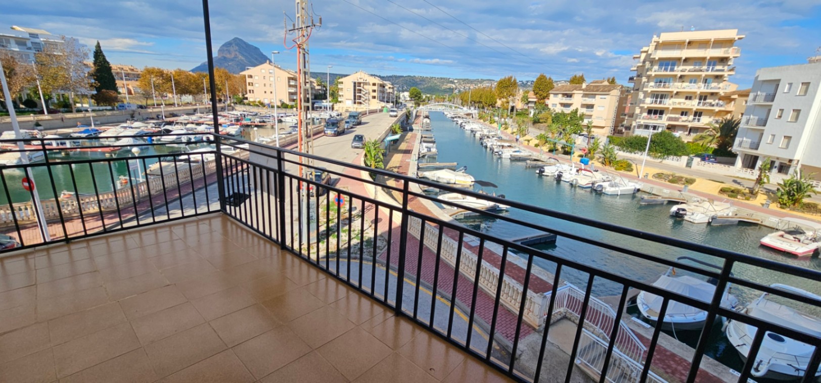 Geflieste Terrasse mit schwarzem Metallgeländer und Blick auf den Jachthafenkanal, die Boote und das charakteristische Montgó-Massiv im Hintergrund.
