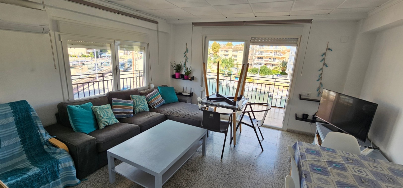 Heller Wohnbereich mit grauem Ecksofa, Klimaanlage und Zugang zur Terrasse mit Blick auf den Kanal und den Jachthafen von Jávea.
