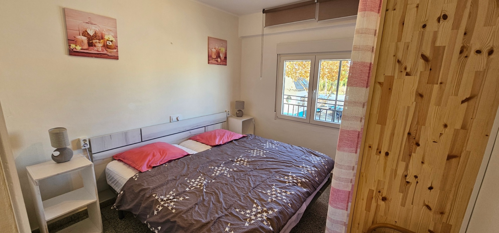 Helles Schlafzimmer mit Doppelbett, hellem Holz-Kopfteil und weißen Nachttischen. Ein Fenster mit Außenansicht sorgt für reichlich natürliches Tageslicht.
