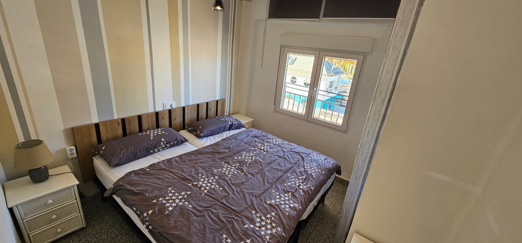 Schlafzimmer mit Doppelbett, Kopfteil aus Holzlamellen und gestreifter Tapete. Das Fenster bietet natürliches Licht und einen Blick auf den nahegelegenen Jachthafen.