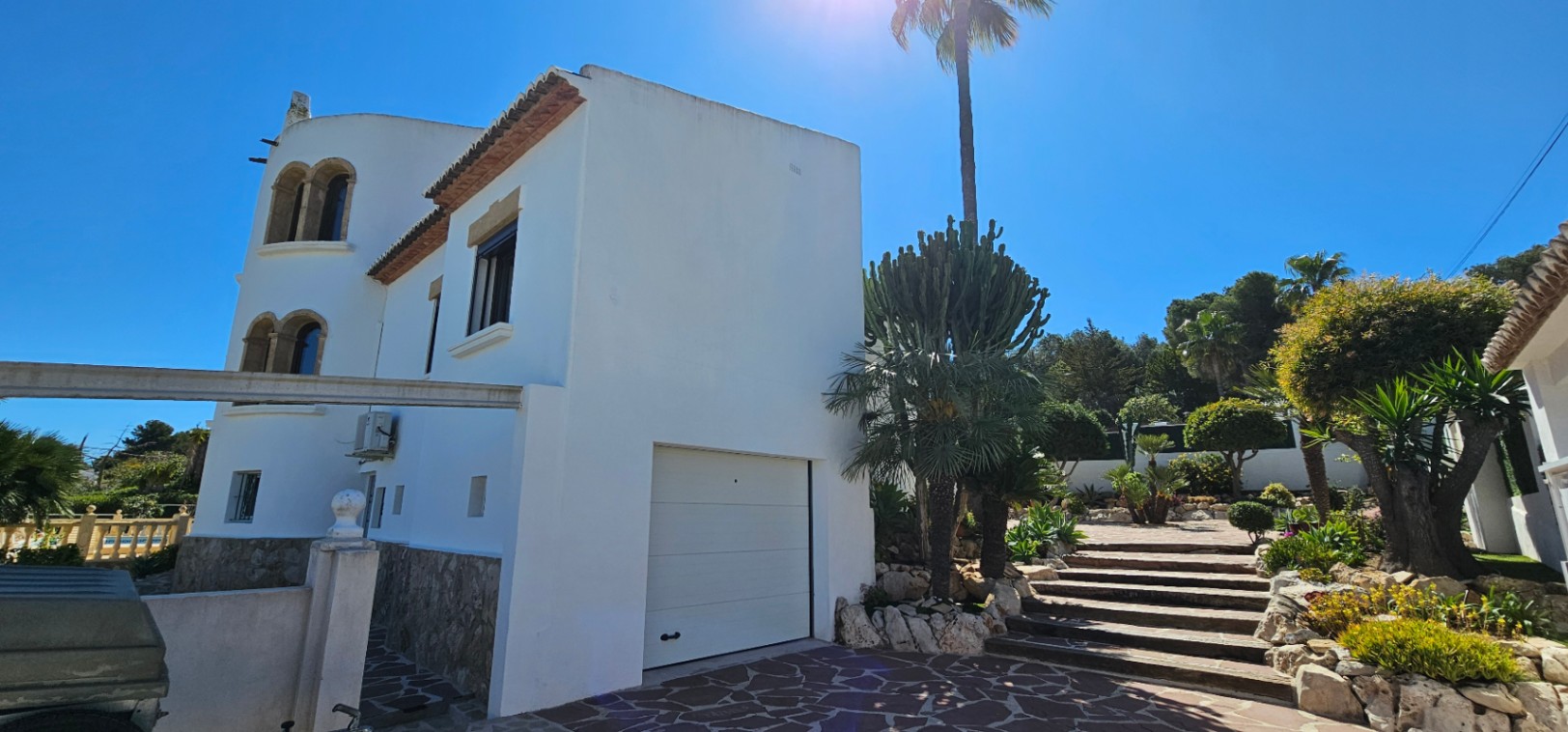 Außenansicht der weißen Villa mit Natursteinsockel, integrierter Garage und einer von mediterranen Pflanzen und Palmen gesäumten Steintreppe.