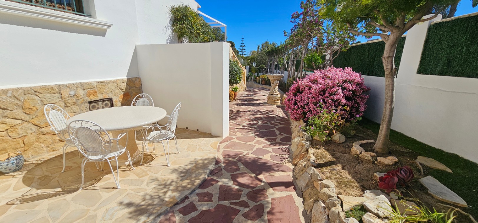 Außenbereich mit Natursteinpflaster, weißen Schmiedeeisenmöbeln und angelegtem Garten mit mediterraner Bepflanzung und blühenden Sträuchern.