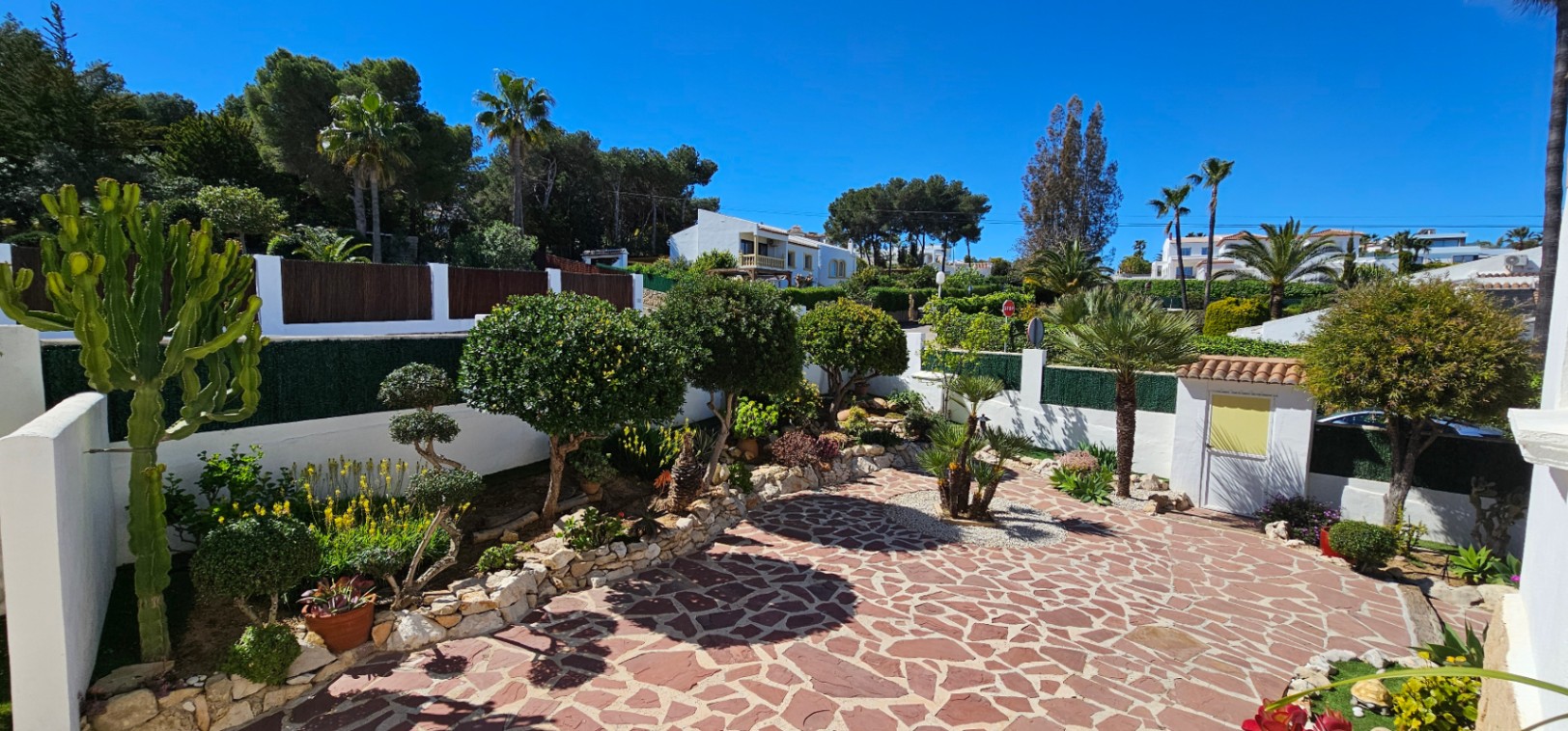 Außenbereich mit unregelmäßiger Natursteinpflasterung, umgeben von Hochbeeten mit mediterranen Pflanzen, Kakteen und Palmen unter blauem Himmel.