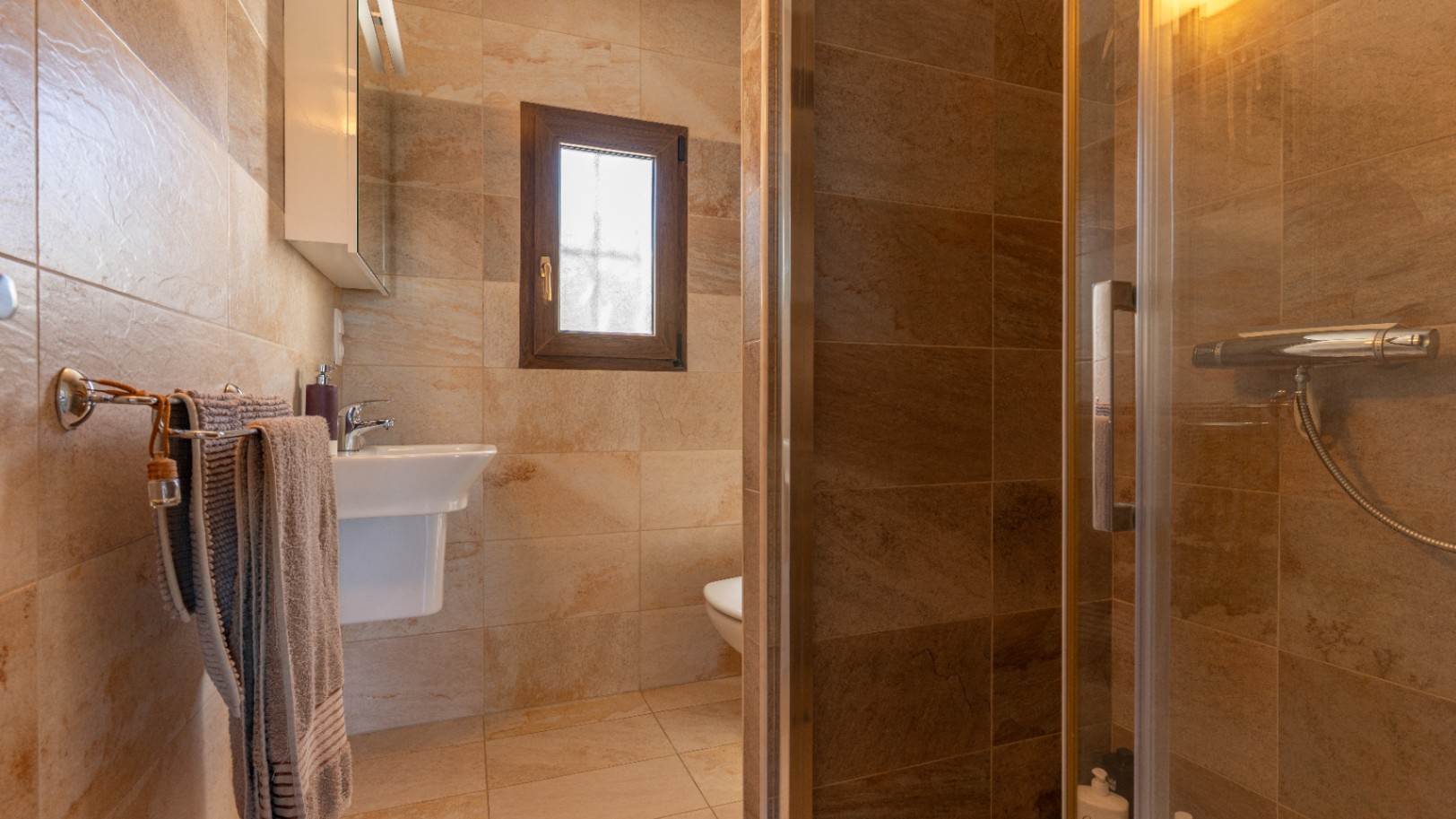 Badezimmer mit erdfarbenen Keramikfliesen, einer Glasduschkabine, einem wandhängenden Waschbecken und einem Holzfenster mit Sichtschutzglas.