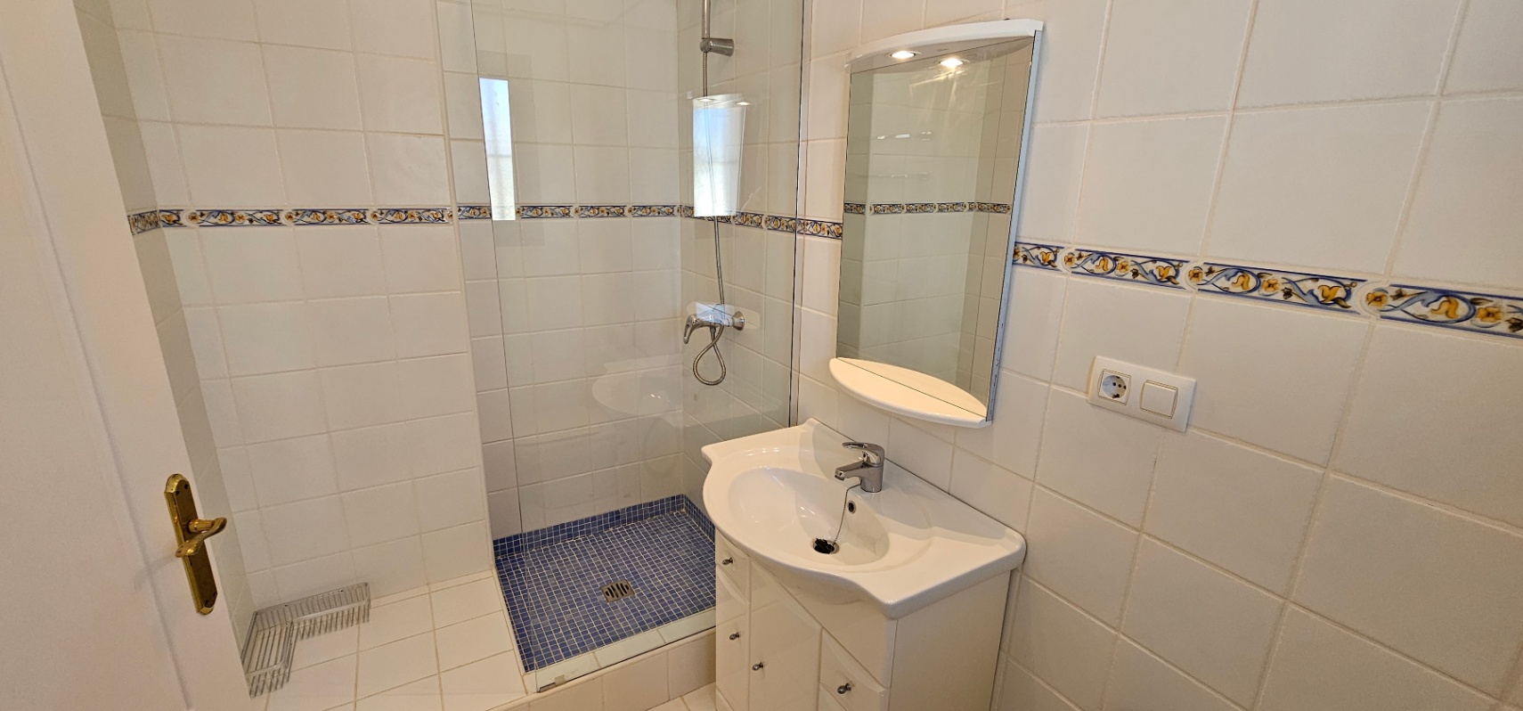 Badezimmer mit Glasduschkabine, blauem Mosaikboden und weiß gefliesten Wänden mit traditioneller Bordüre, ausgestattet mit einem weißen Waschtisch.