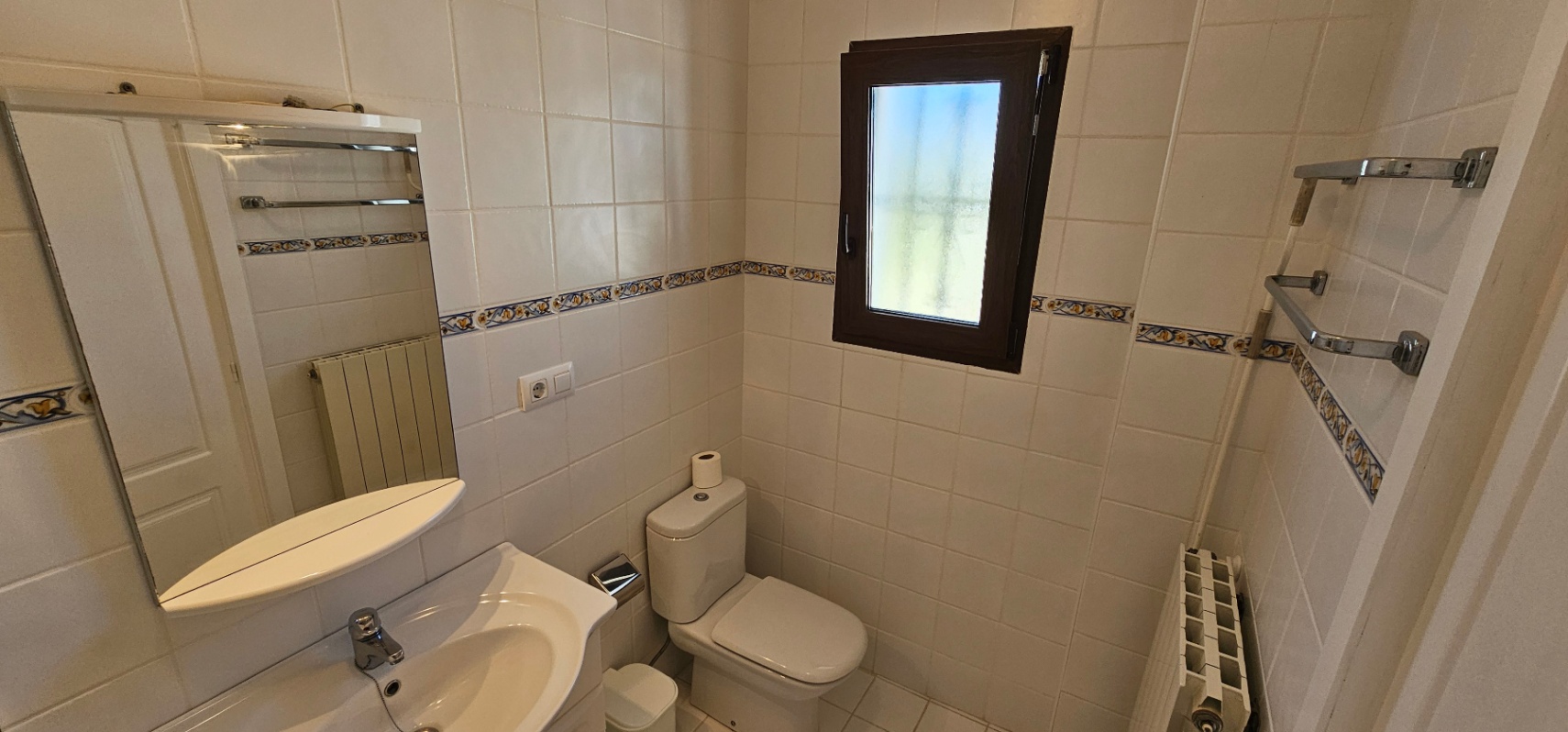 Badezimmer mit weißen Wandfliesen und dekorativer Bordüre, ausgestattet mit Toilette, Waschbecken mit Spiegel und einem dunklen Fensterrahmen.