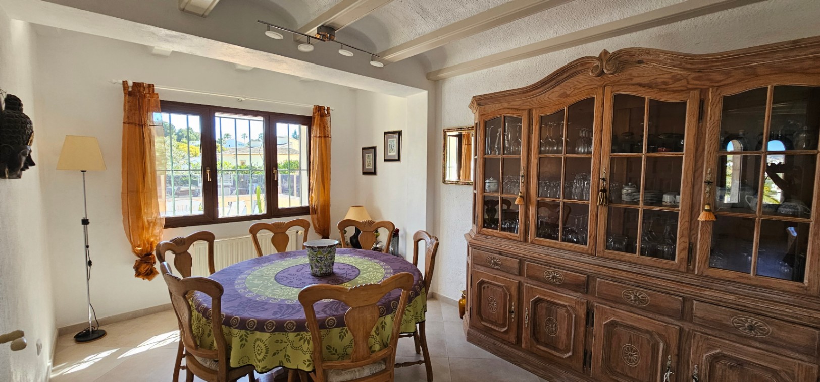 Esszimmer mit rundem Tisch, geschnitzten Holzstühlen und einer großen klassischen Vitrine. Mit Sichtbalken an der Decke und natürlichem Licht durch das Fenster.