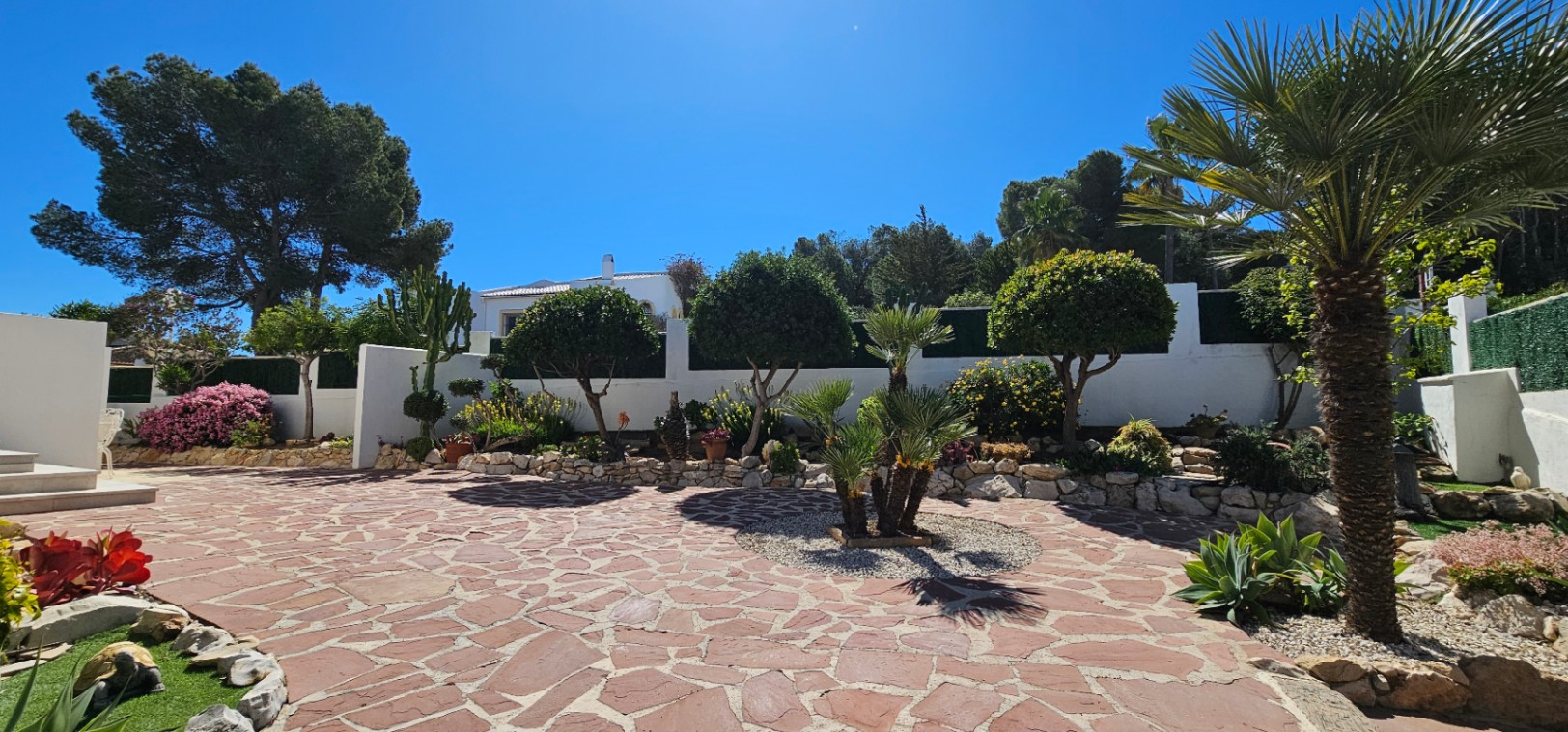Großzügiger Außenhof mit unregelmäßiger rötlicher Steinpflasterung, umgeben von gepflegten Gartenbeeten mit mediterranen Pflanzen, Palmen und weißen Mauern.