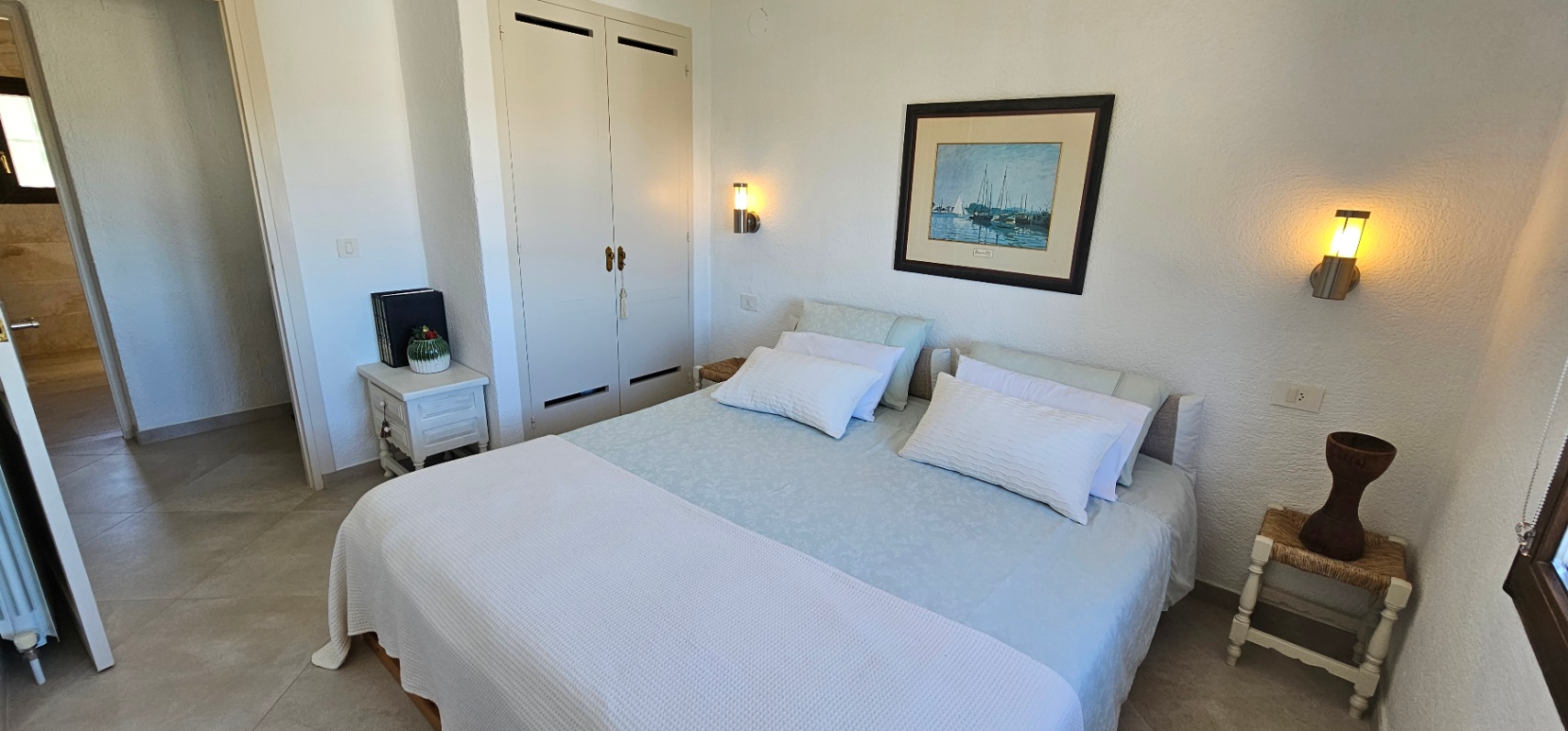 Hell eingerichtetes Schlafzimmer mit Doppelbett, weißen Einbauschränken und Fliesenboden. Wandleuchten sorgen für eine warme Atmosphäre im mediterranen Stil.
