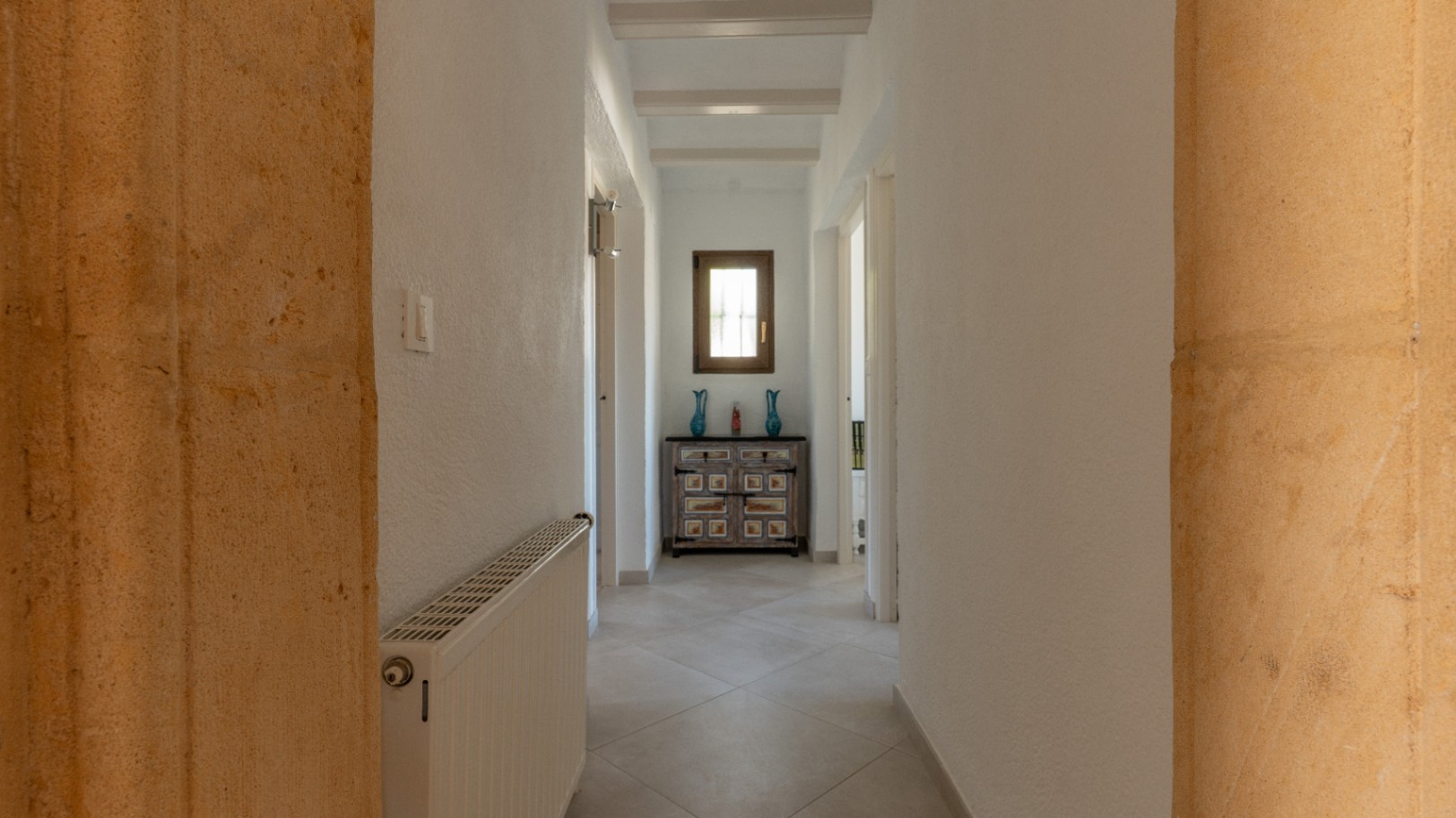 Heller Flur mit weißen Deckenbalken und Tosca-Natursteinbögen. Ausgestattet mit Zentralheizung und einer dekorativen Kommode unter einem Holzfenster.