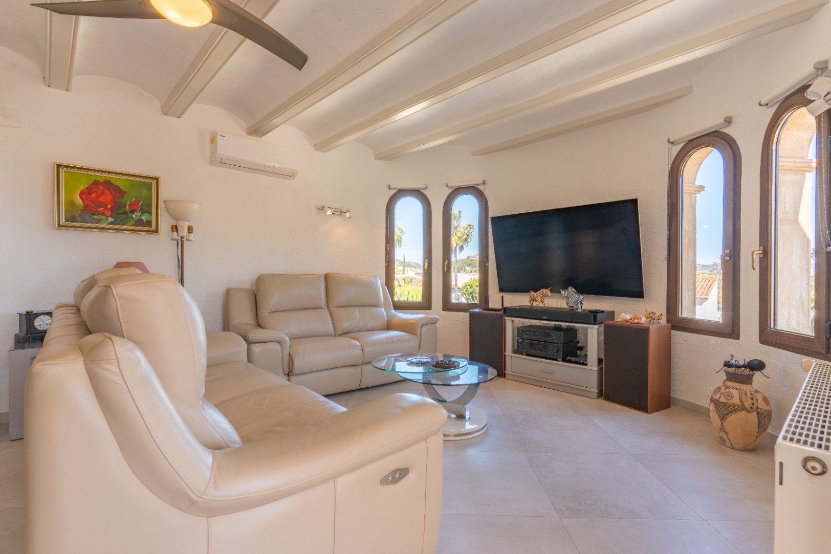 Heller Wohnbereich mit Sichtbalken, cremefarbenen Ledersofas und Rundbogenfenstern. Inklusive Klimaanlage, Deckenventilator und einer modernen Medienecke.