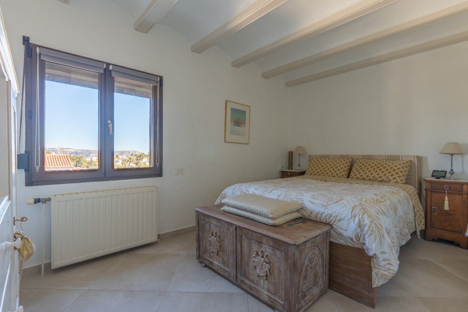 Helles Schlafzimmer mit Sichtbalken an der Decke, dunklem Holzfenster mit Weitblick und einer rustikalen Holztruhe am Fußende des Bettes.