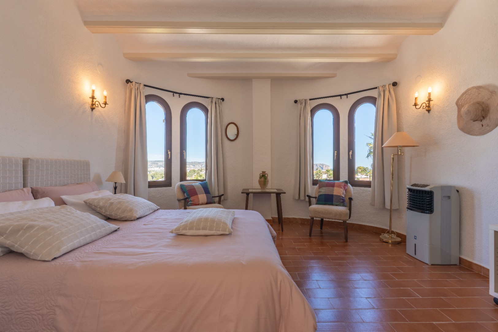 Helles Schlafzimmer mit Sichtbalken, Terrakottaboden und Rundbogenfenstern mit Blick auf Jávea, ergänzt durch klassische Wandleuchten im Kerzenstil.