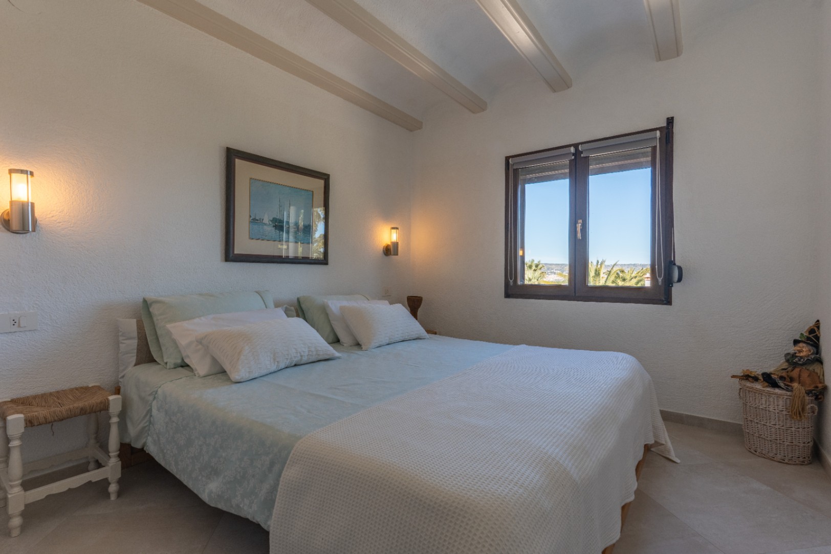 Helles Schlafzimmer mit sichtbaren Deckenbalken, modernen Wandleuchten und einem Holzfenster mit Blick auf Palmen und die umliegende Landschaft.
