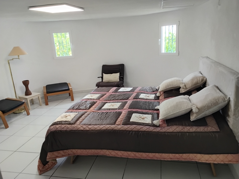 Helles Schlafzimmer mit weißen Fliesenböden, einem Doppelbett mit gepolstertem Kopfteil und zwei Fenstern mit Sicherheitsgittern für natürliches Tageslicht.