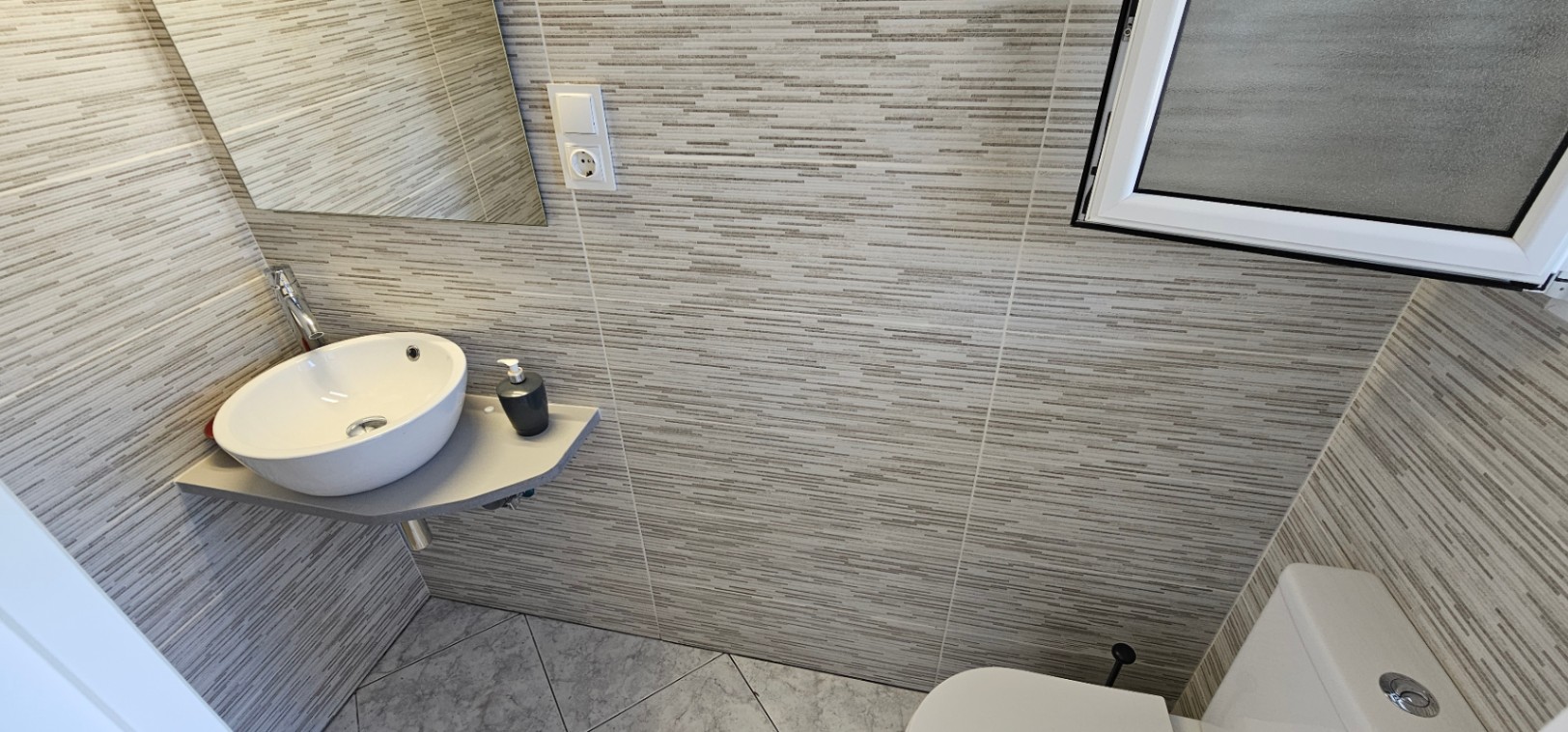 Kompaktes Gäste-WC mit Eck-Aufsatzwaschbecken, horizontal gestreiften grauen Wandfliesen und diagonal verlegten Bodenfliesen.