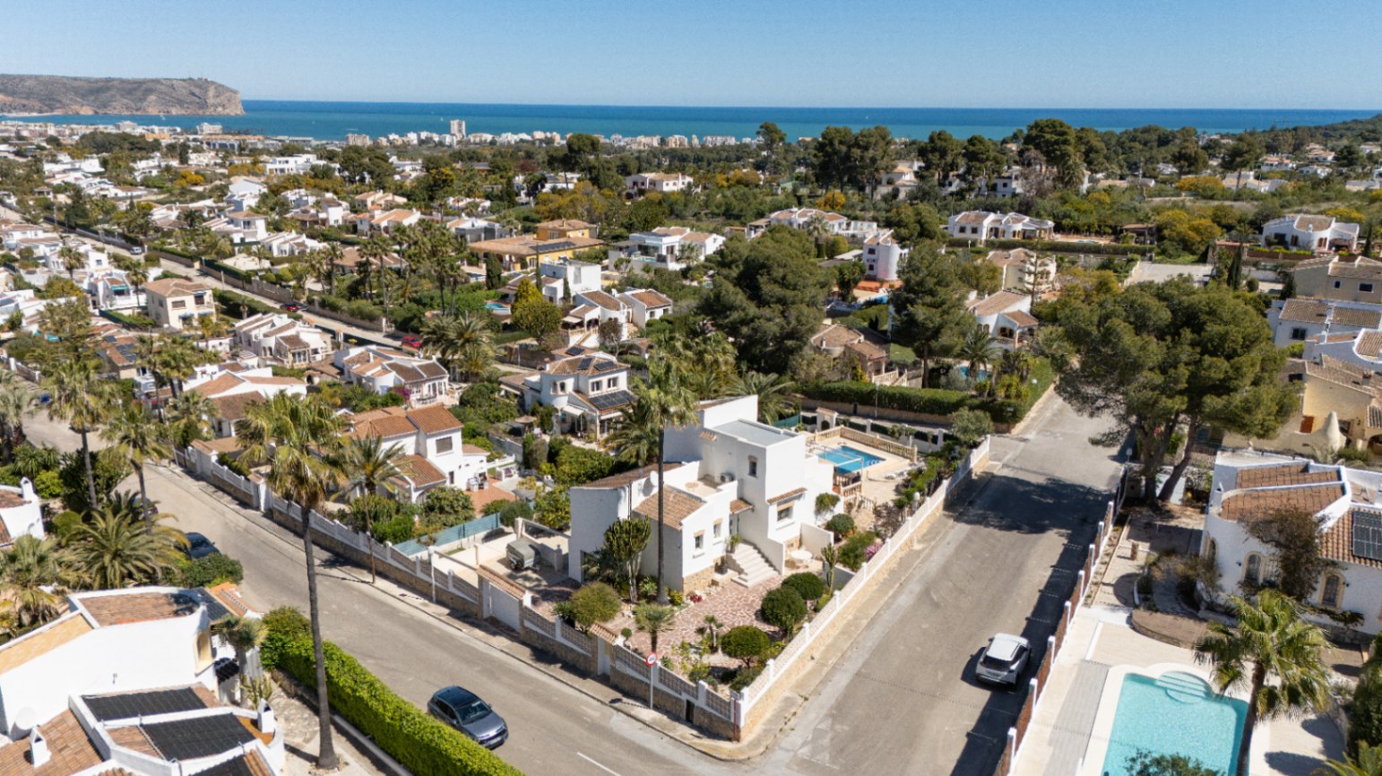 Luftaufnahme der weißen Villa auf einem Eckgrundstück in Pinosol, Jávea. Gezeigt werden der private Pool, der gepflegte Garten und der weite Blick auf das Mittelmeer am Horizont.