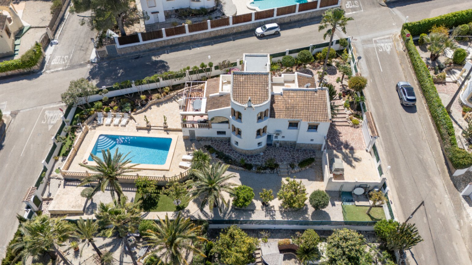 Luftaufnahme des Anwesens auf einem Eckgrundstück mit rechteckigem Pool, markantem Rundturm und mediterranem Garten mit Palmen.