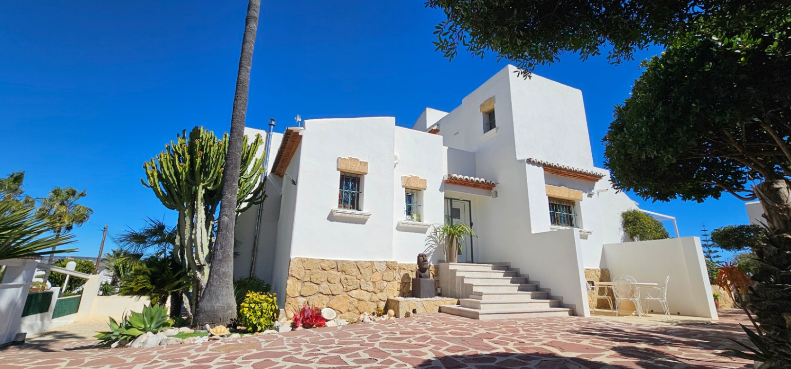 Mediterrane Fassade mit weißen Wänden, Natursteinsockel und Treppenaufgang. Der Außenbereich umfasst eine gepflasterte Terrasse mit Kakteen und Palmen.