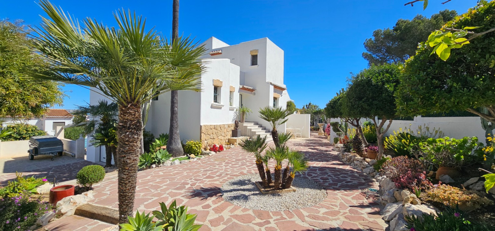 Mediterrane Villa mit weißer Fassade und Natursteinsockel, eingebettet in einen gepflegten Garten mit Palmen und gepflastertem Steinweg.
