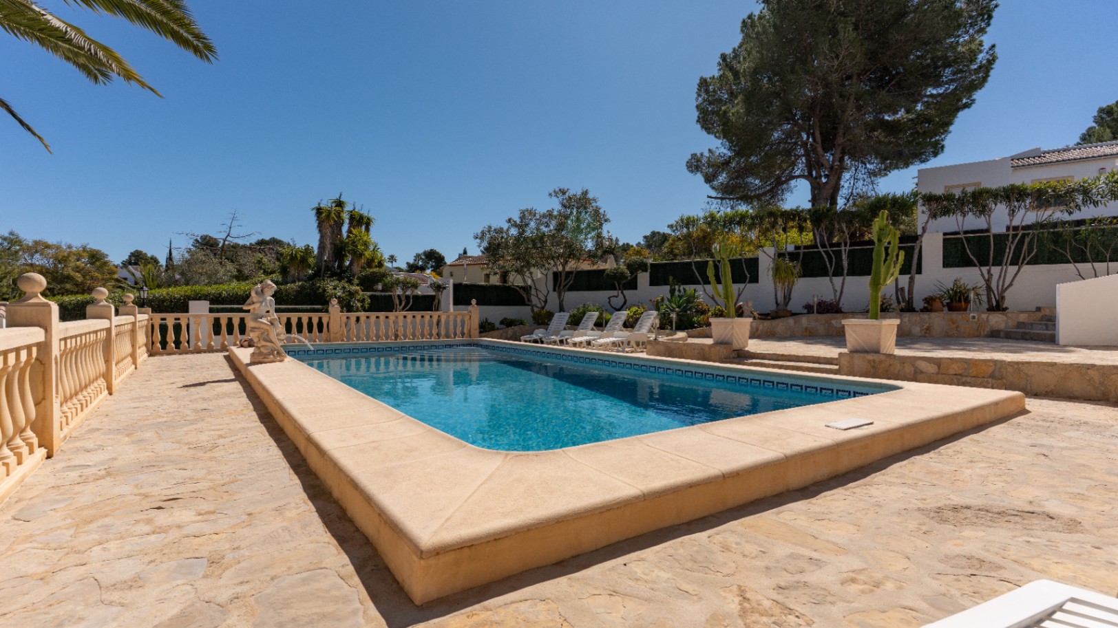 Rechteckiger Pool mit Natursteinterrasse, klassischer Balustrade und dekorativer Statue, umgeben von mediterraner Bepflanzung und schattenspendenden Pinien.