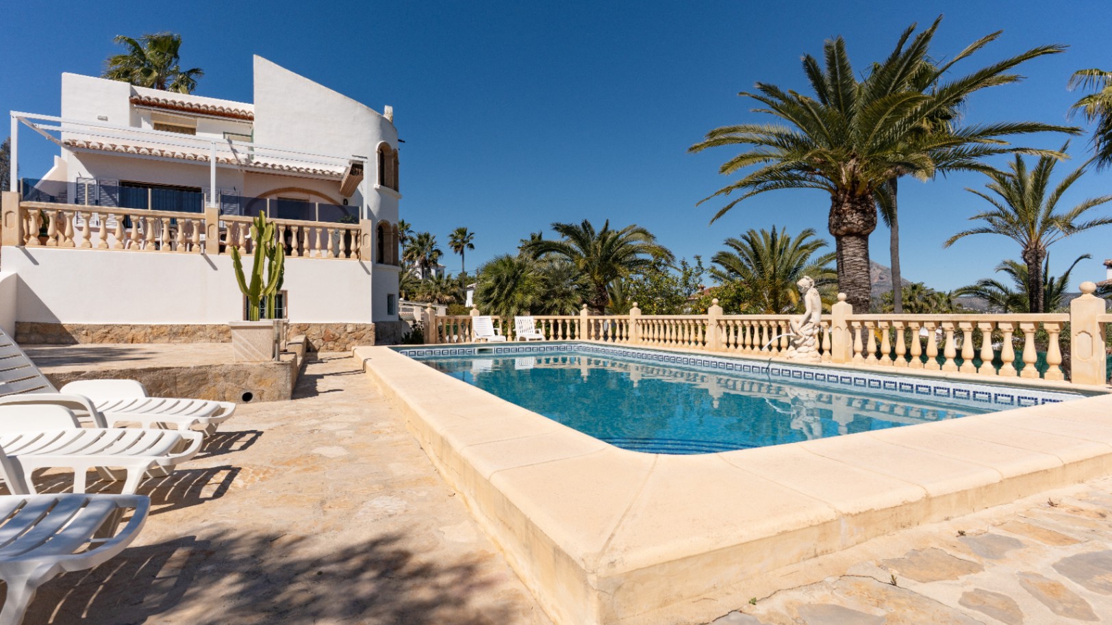 Rechteckiger Pool mit weitläufiger Steinterrasse und Sonnenliegen, umgeben von Palmen und der weißen mediterranen Fassade mit traditionellen Balustraden.