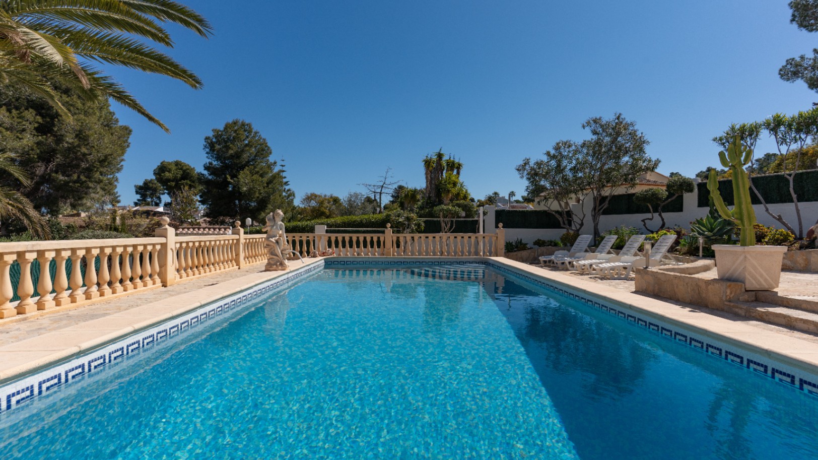 Rechteckiger Swimmingpool mit blauen Mosaikfliesen, umgeben von einer Steinterrasse mit klassischer Balustrade, Sonnenliegen und mediterraner Bepflanzung.