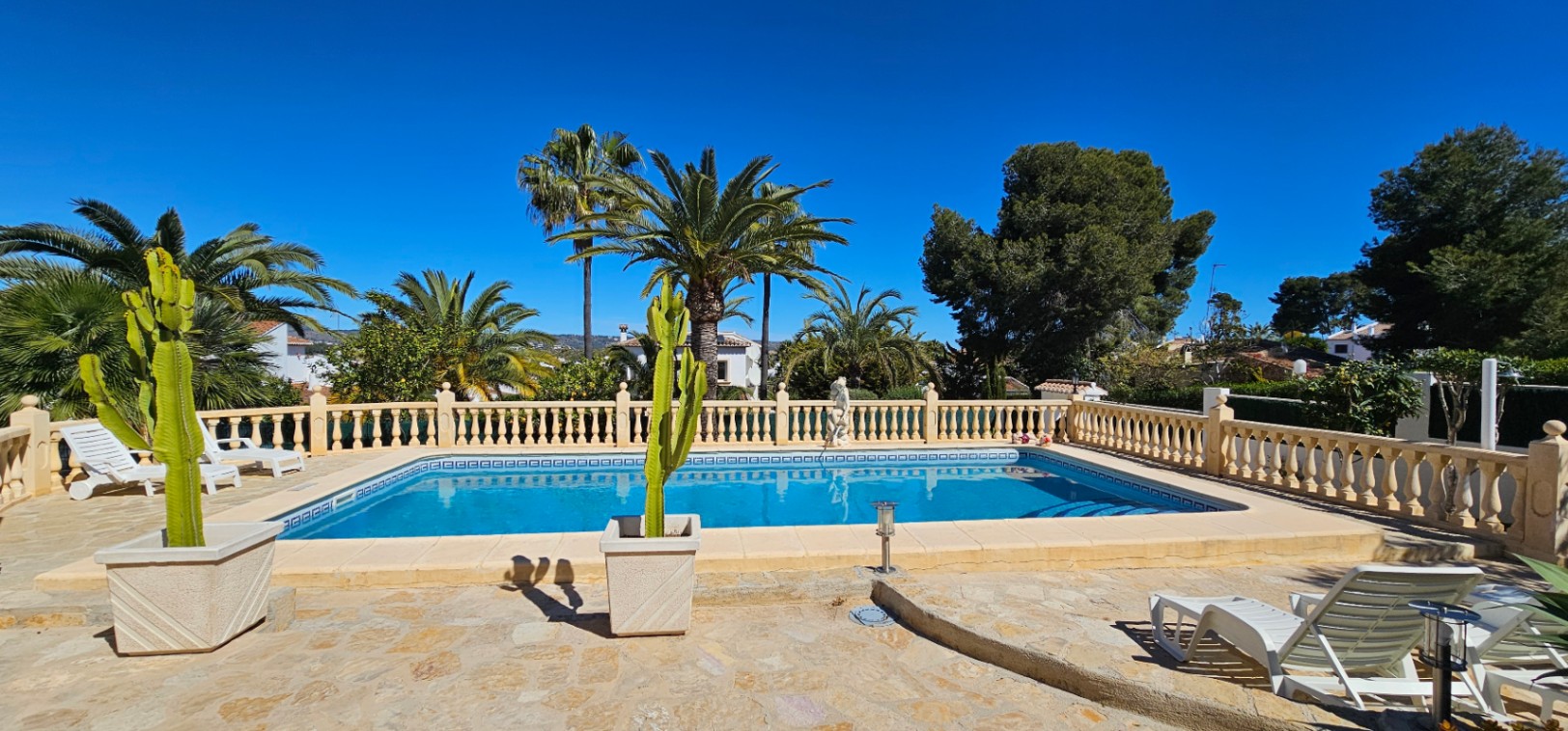 Rechteckiger Swimmingpool mit großzügiger Natursteinterrasse, klassischer Balustrade, ausgewachsenen Palmen und dekorativen Kakteen in Kübeln.