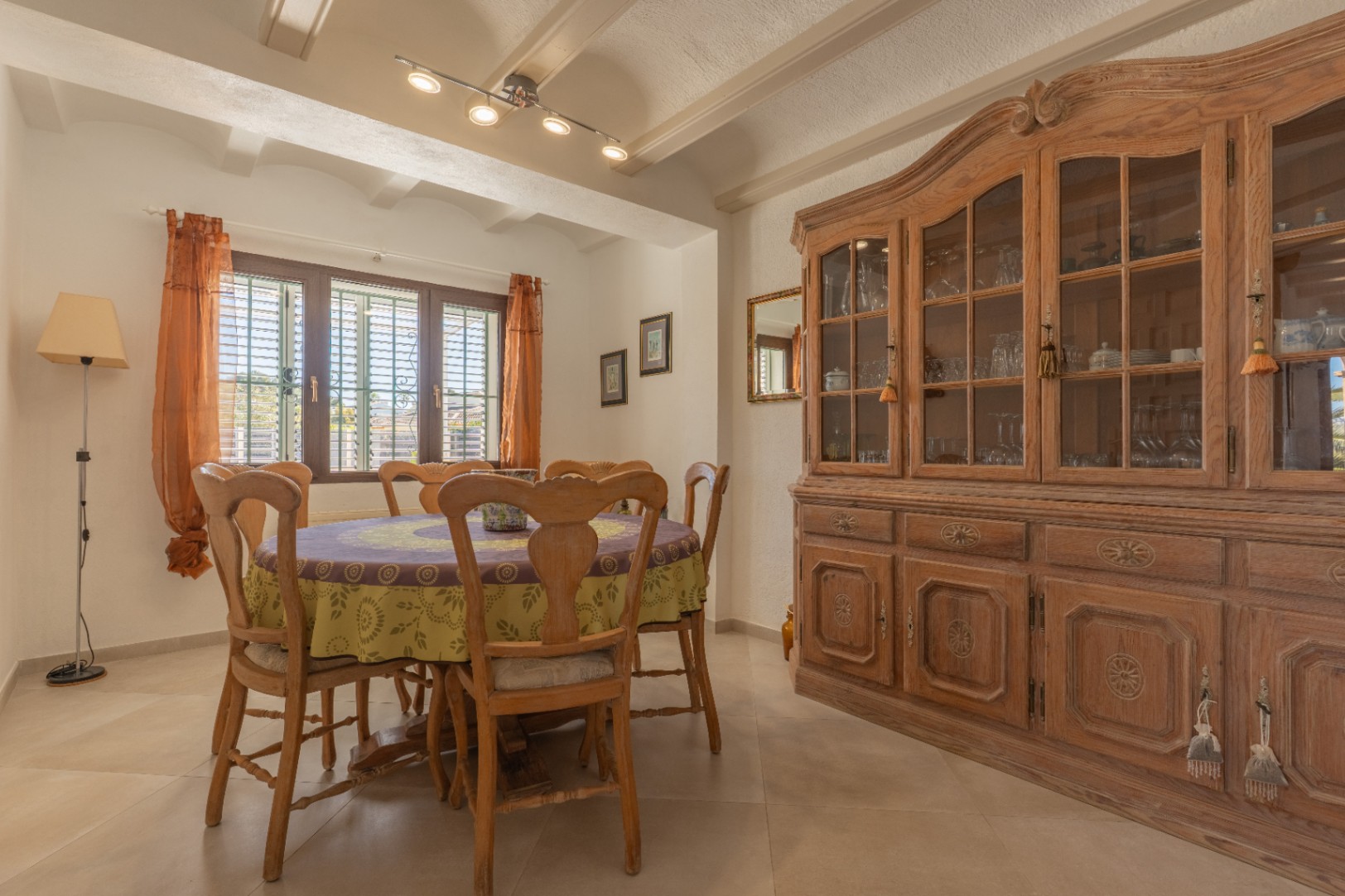 Rustikaler Essbereich mit Sichtbalkendecke, rundem Holztisch und einem großen klassischen Buffetschrank. Ein Fenster mit Fensterläden sorgt für Tageslicht.