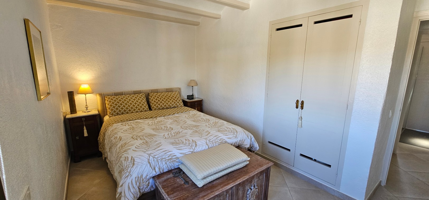Schlafzimmer mit sichtbaren Deckenbalken, Einbauschrank mit weißen Türen und einer rustikalen Holztruhe am Fußende des Bettes.