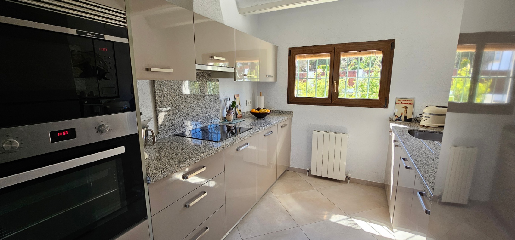 Separate Küche mit Hochglanzfronten, Granitarbeitsplatte und integrierten Geräten. Ein Holzfenster sorgt für natürliches Licht und Blick in den Außenbereich.