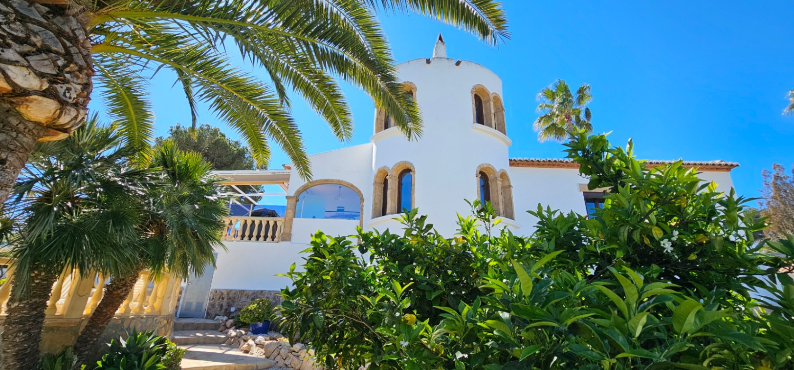 Weiße Villa mit markantem Rundturm und Rundbogenfenstern aus Tosca-Stein, eingebettet in einen mediterranen Garten mit Palmen und Zitrusbäumen.