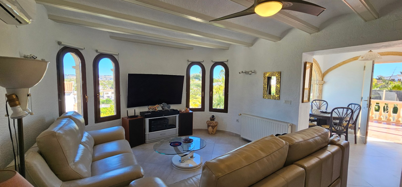 Wohnbereich mit Ledersofas, weißen Balkendecken und Rundbogenfenstern. Der Raum umfasst eine Medienecke und bietet Zugang zum Esszimmer durch einen Rundbogen.